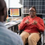 African businesswoman sitting on wheelchair and discussing work with businessman while they sitting opposite the table with computer at office