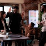 Female manager in wheelchair and group of people laughing on job break, having fun in disability friendly office. Woman with chronic health condition chatting with cheerful colleagues.