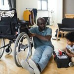 African little boy sitting on floor in the room with tool box and helping his father with disability to fix wheelchair